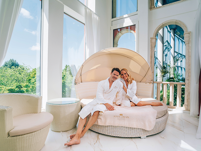 A couple is sitting on a large white lounger, with palm trees visible in the background.