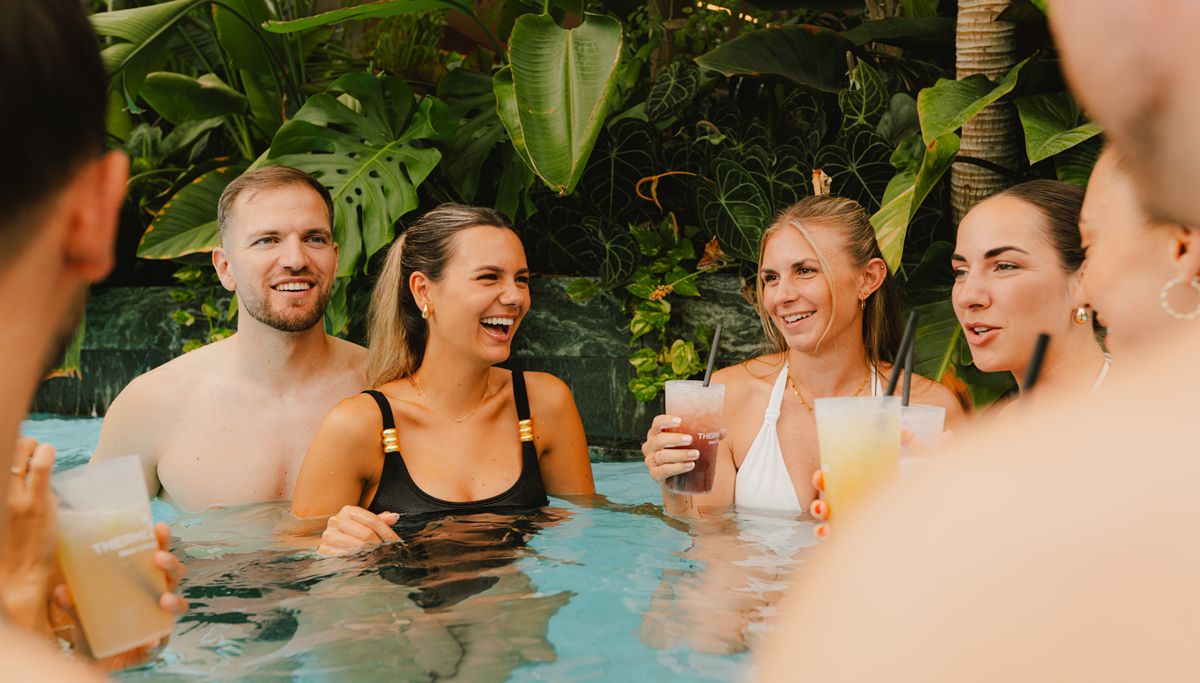 Eine Freundesgruppe entspannt zusammen im Pool mit Cocktails in der Hand.