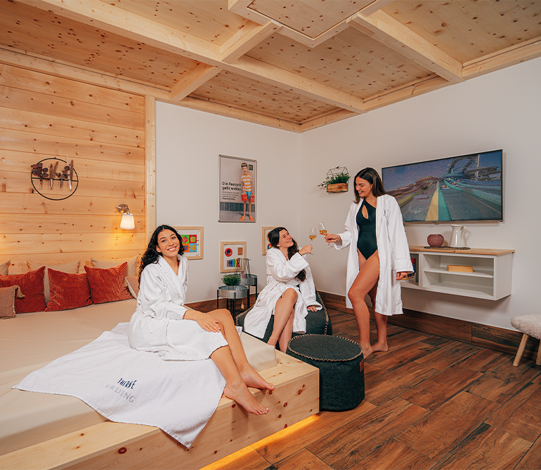 Drei Frauen entspannen in einem urig modernen Zimmer mit einem großen Bett in Holzoptik mit Kissen, Decke und Sitzkissen.