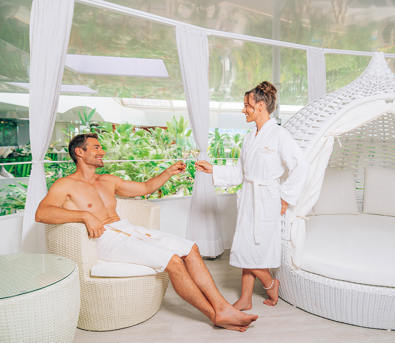 A couple toasts with champagne. A bright, cheerful, white setting. Quality time together at the spa. 