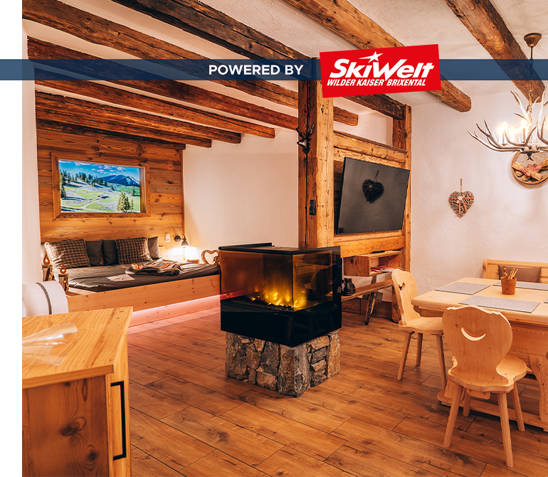 Raum in uriger Almhütten-Atmosphäre mit Tisch, Stühlen, Fernseher, großem Bett und Effektfeuer.