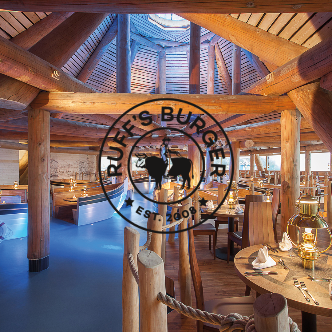 Innenraum eines Restaurants mit rustikaler Holzkonstruktion, runden Tischen mit Lampen und Sitzbänken, blauem Boden und Dachfenster