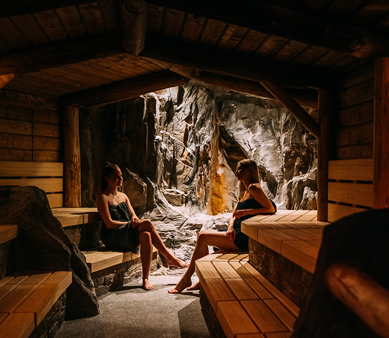 Two women in bathrobes are sitting in a wood-paneled saltwater tunnel with rock walls at the Erding Thermal Baths