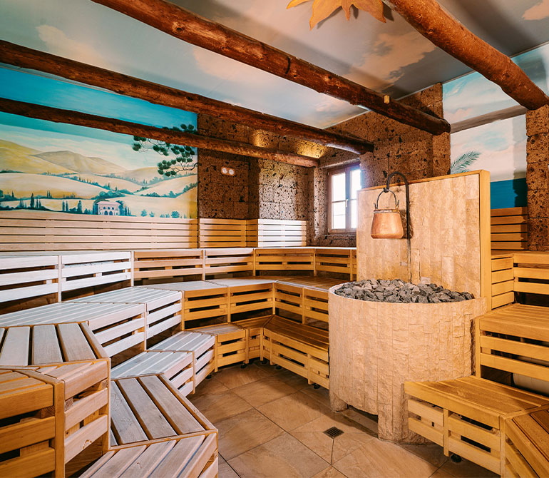 Sauna room with light-colored wooden benches, a stone stove with a kettle, and a mural depicting a Tuscan landscape