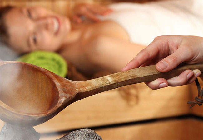 A hand holds a wooden spoon over hot stones in a sauna; in the background, a woman lies relaxed