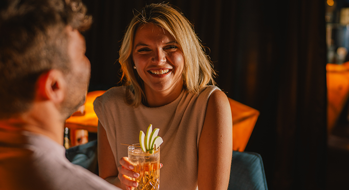 Frau sitzt mit Cocktail an Bar und lächelt in die Kamera