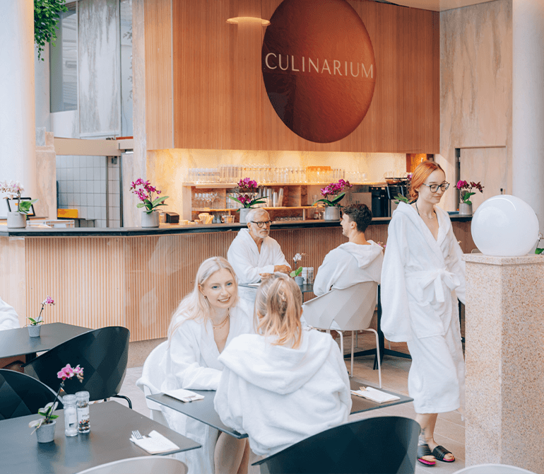 Sitzbereich mit Tischen und Stühlen neben einem Pool unter einer großen Glaskuppel mit tropischen Pflanzen im Thermenrestaurant