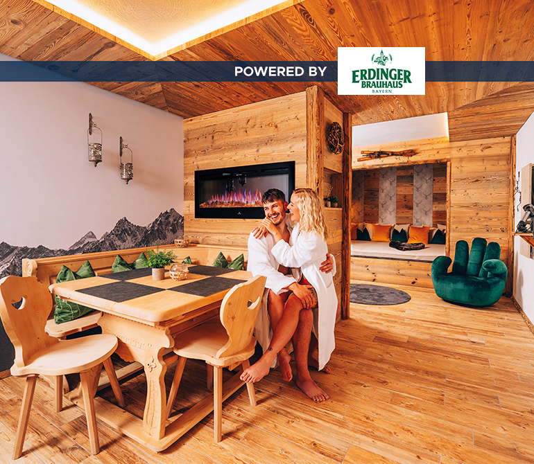 Ein Paar sitzt an einer großen Eckbanksitzgruppe aus Holz in einem Raum im Hüttenstil mit Effektfeuer im Hintergrund.