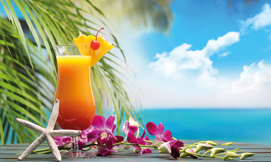 Fruity cocktail with pineapple and cherry on a tropically decorated table with orchids, starfish and a view of the sea under a blue sky.