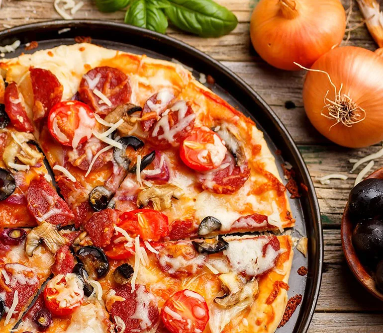 Pizza mit Salami, Kirschtomaten, Oliven und Käse auf einem schwarzen Teller, umgeben von Zwiebeln und Basilikum auf Holztisch