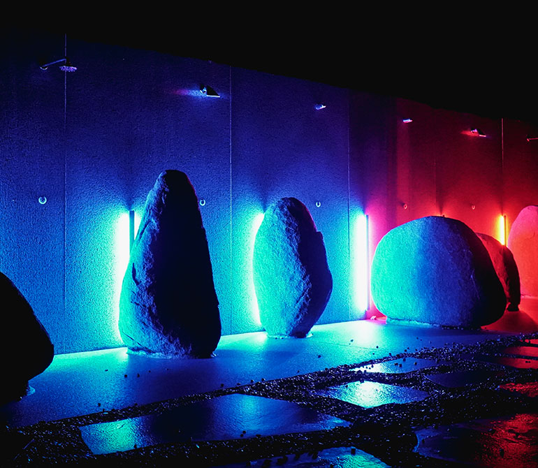 Stone objects arranged in a row in front of a wall with blue and red neon lights, inspired by Stonehenge, at the Therme Erding