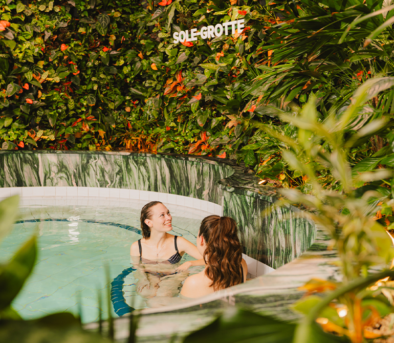 Two women are relaxing in a round pool surrounded by green living walls.
