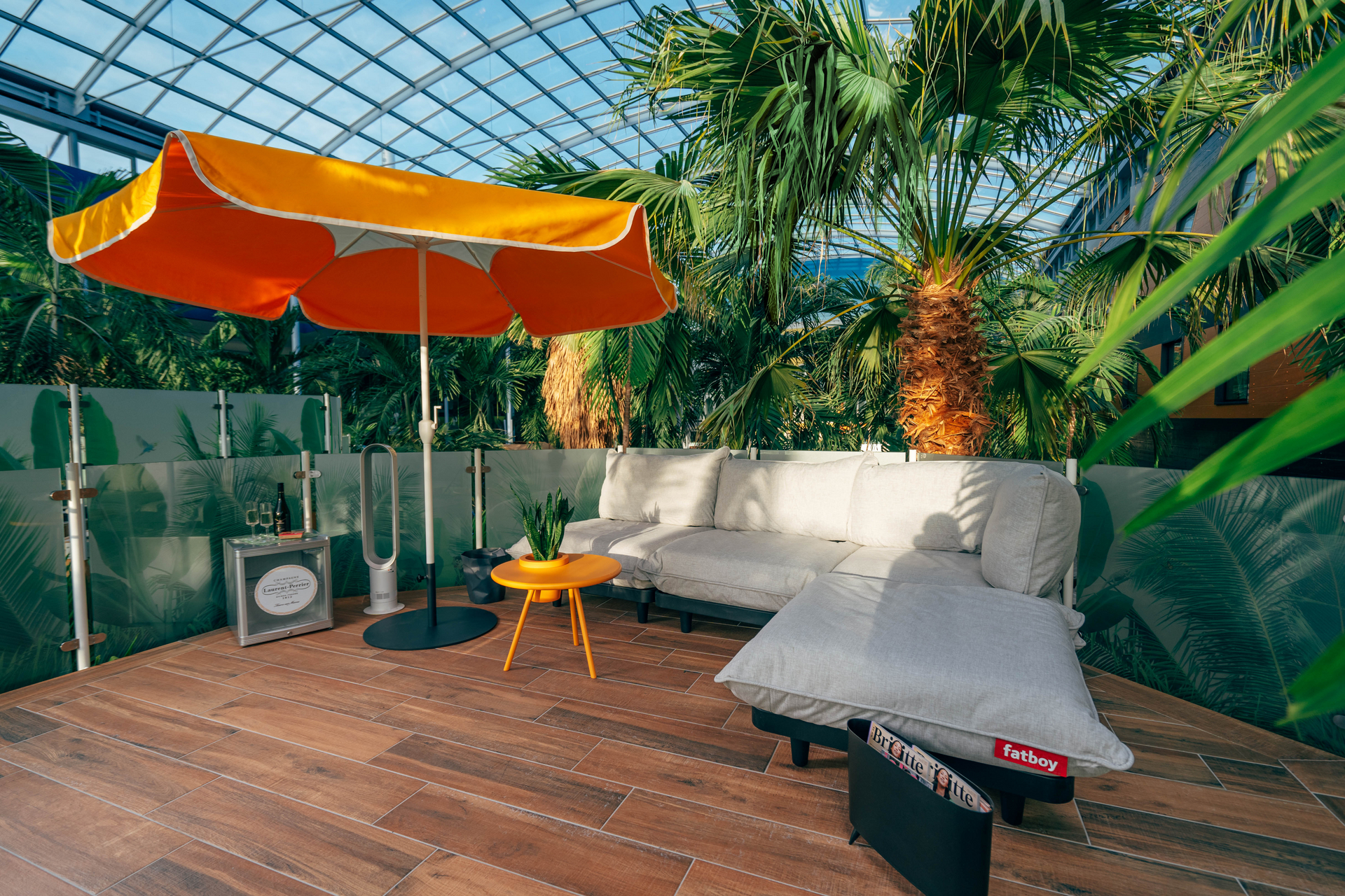 Stilvoller, erhöhter Lounge-Bereich mit Couch, Schirm, Kühlschrank und Palmen.