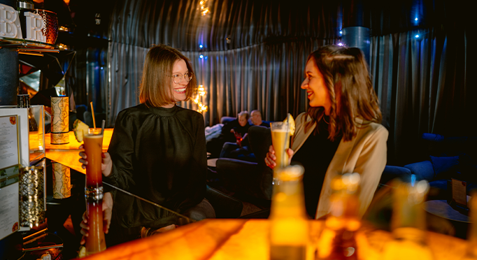 Two friends stand in the illuminated Kaleidoscope Bar and toast with a cocktail