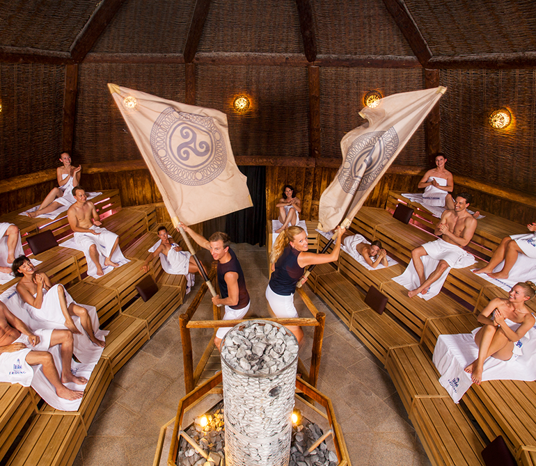 People in a round sauna sit on wooden benches; two people wave large flags bearing Celtic symbols over a central pile of stones