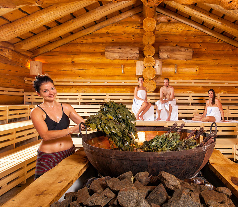 Three people in a wooden sauna room; two are sitting on benches with white towels, and a woman is waving a flag next to a large wood-burning stove decorated with green birch branches