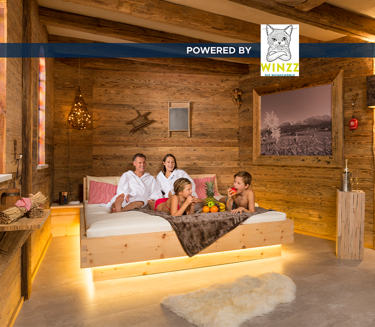 Eine Familie mit zwei Kindern liegt auf einem großen Bett in einem Raum im Holz-Hüttenstil mit bunten Fenstern.