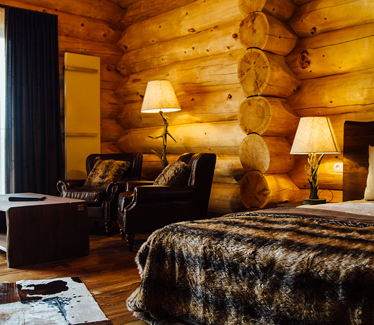 Rustic chalet room with wooden walls, a double bed, a deer head on the wall, leather armchairs, a wooden table, and an antler lamp