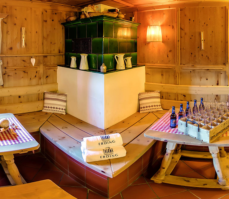 A cozy room with green tiled radiators, wooden walls, wooden benches with cushions, tables covered with checkered tablecloths, beer bottles and glasses, and two white towels printed with “Therme Erding”