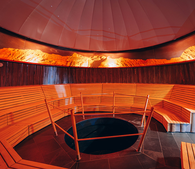 A wooden bench in a circular room with warm orange lights and an opening in the center of the floor