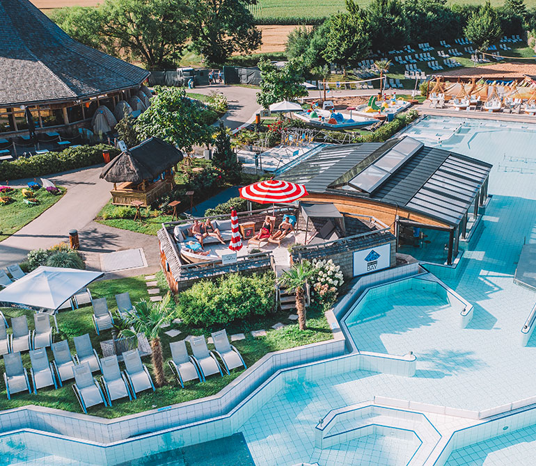 Große eingefasste Outdoor-Lounge mit Sonnenschirm, umgeben von Pool und grüner Liegewiese.