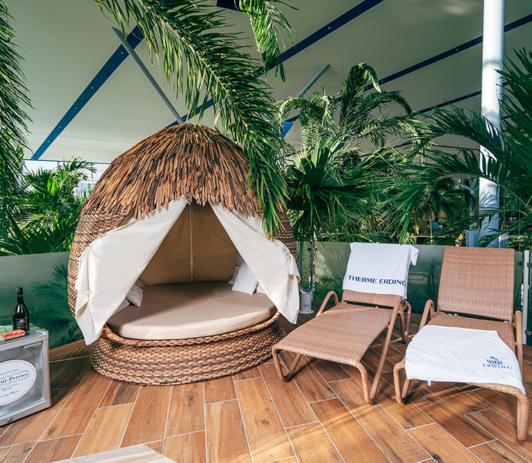 Eine Liege aus Rattan in Form einer Kokosnuss steht neben einer weiteren Liege in stilvoller, umzäunter Palmenlounge.