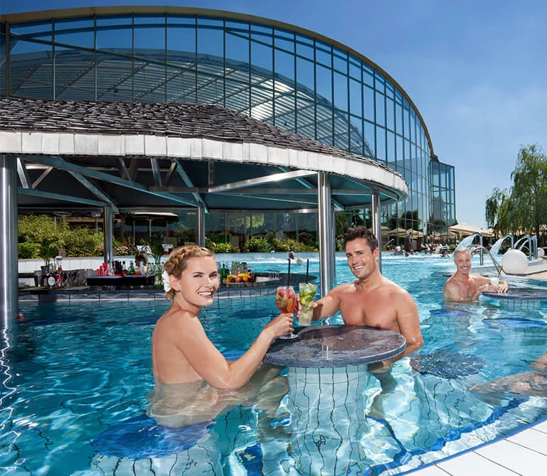 Zwei Personen im Außenpool der Therme Erding, zwei stoßen mit Cocktails an, moderne Glasarchitektur im Hintergrund