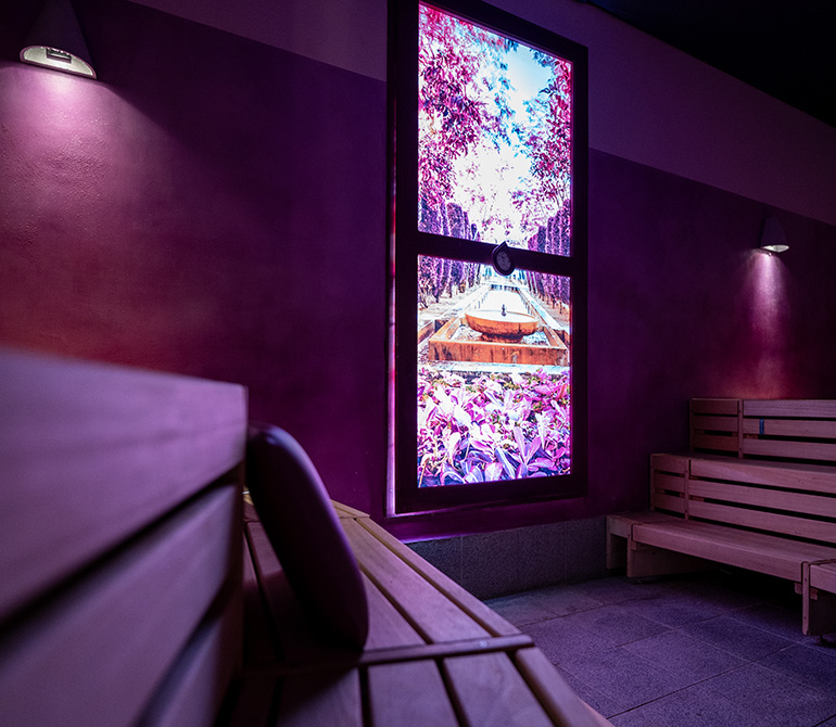 Sauna room with wooden benches and an illuminated window featuring a nature scene in shades of purple