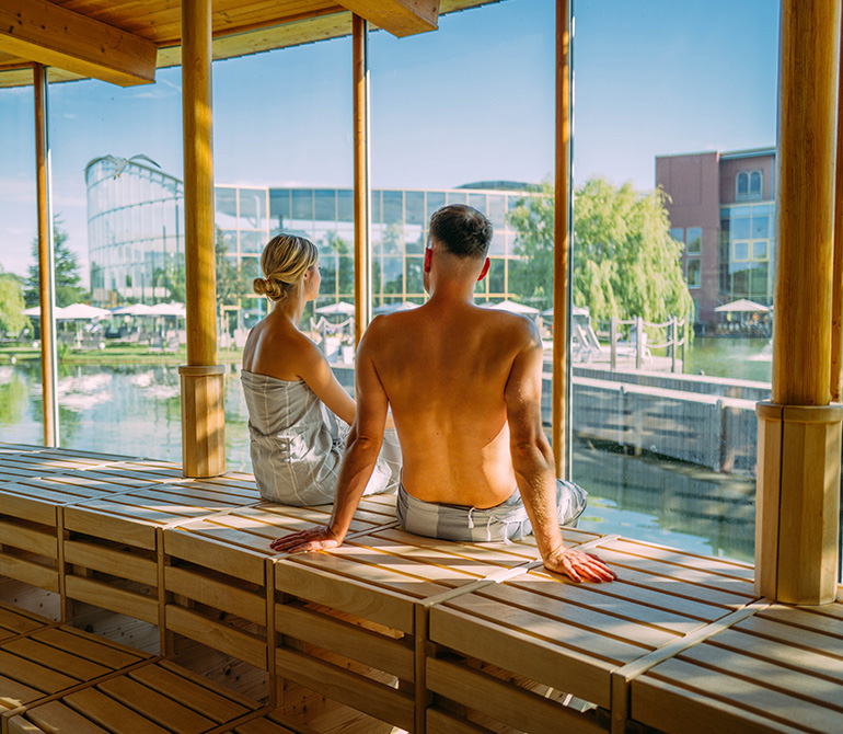 Two people in bathrobes are sitting in a sauna with large windows overlooking a lake and modern buildings