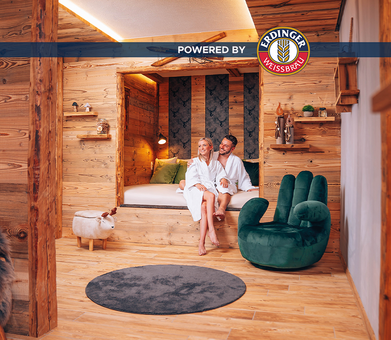 Ein Paar sitzt in einem großen Raum im Hüttenflair auf einem Bett.